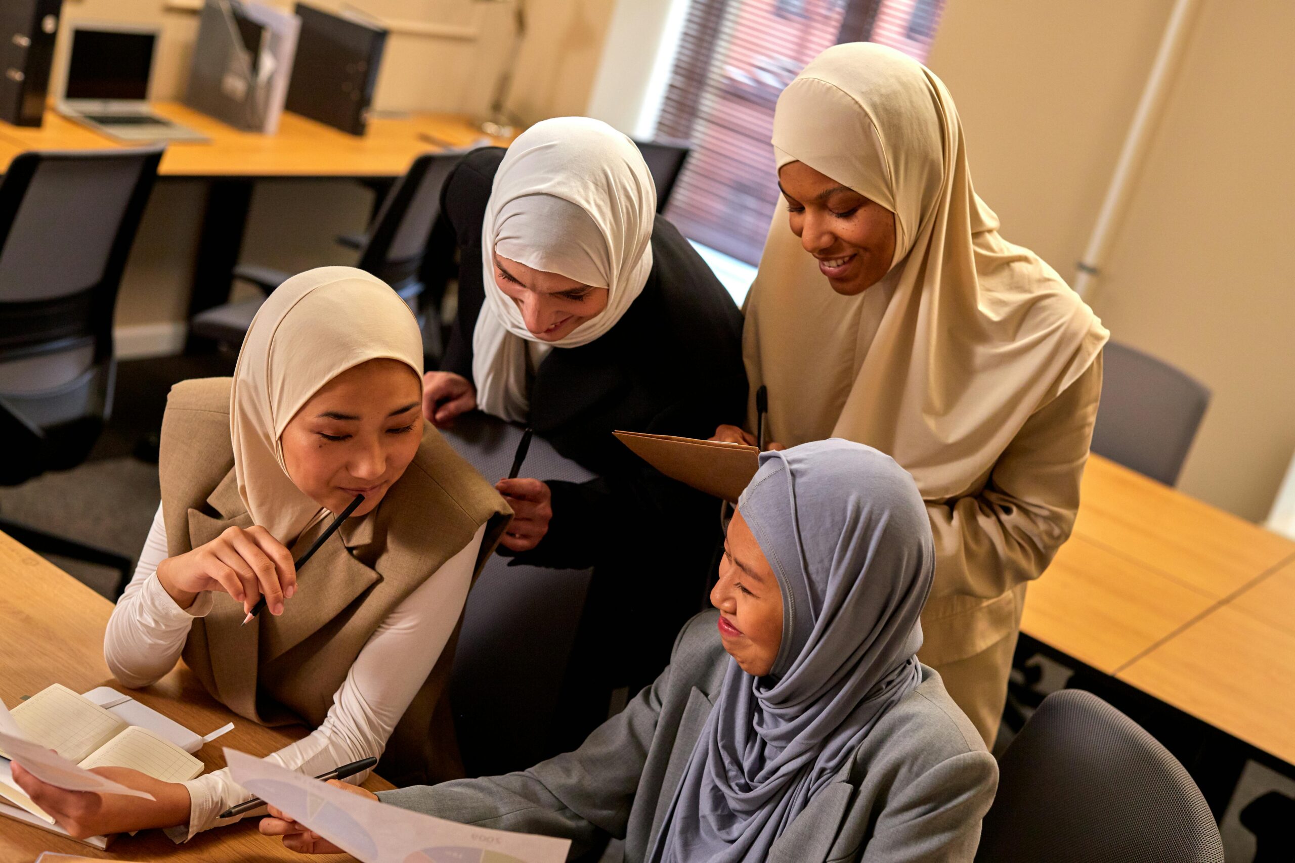 Four diverse women in hijabs engaged in a lively discussion around work documents in a modern office.