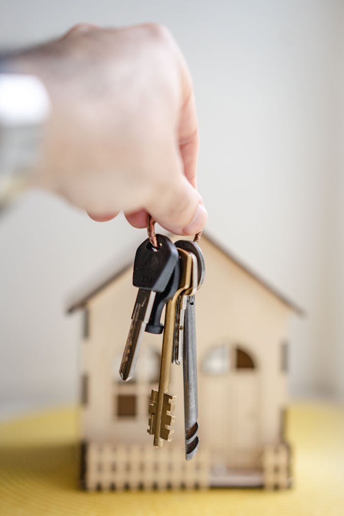 about-01 Close-up of a hand holding keys with a miniature wooden house in the background, symbolizing real estate investment.